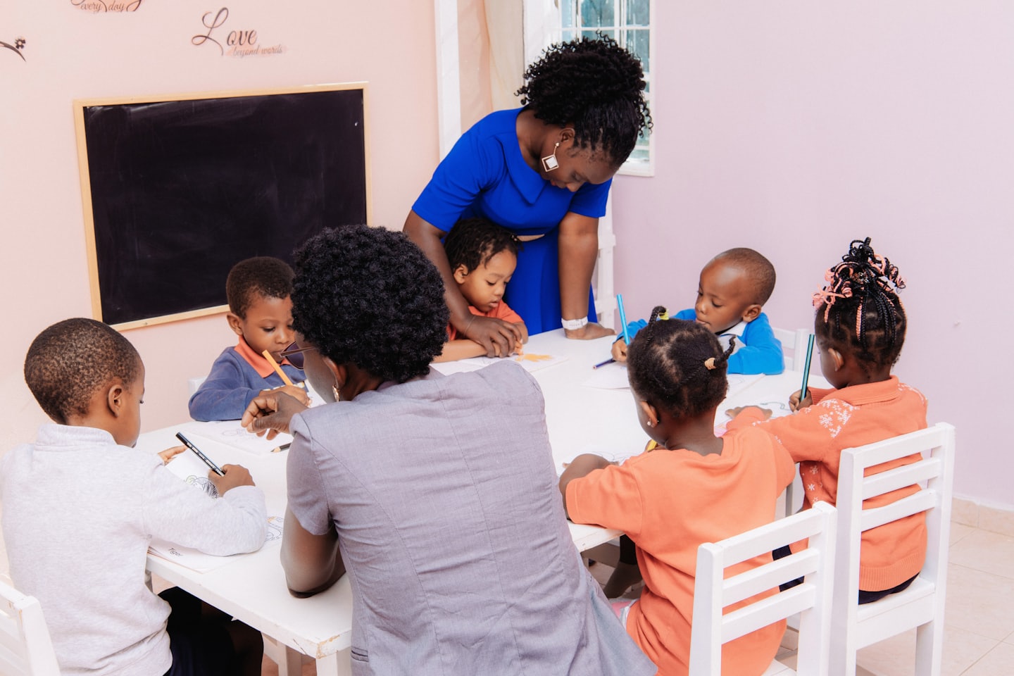 Woman helps boy draw at table with other children coloring.