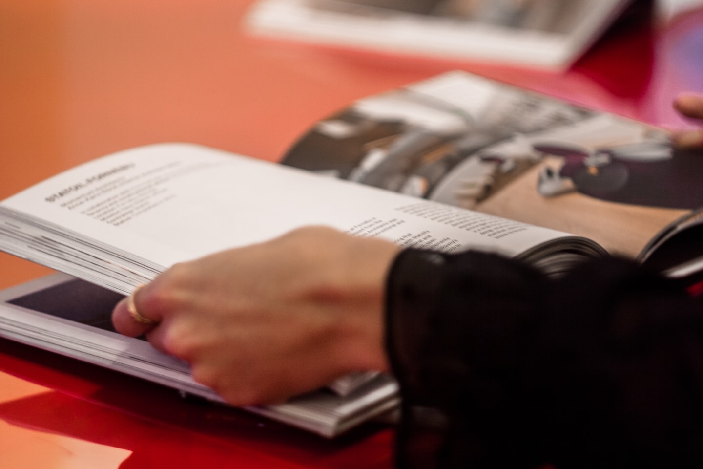 Hand with gold ring turns pages of a book on red table.